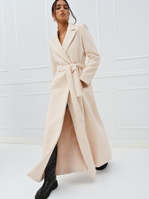 Lungo cappotto beige realizzato in tessuto morbido, con vita con cintura, collo a revers e tasche laterali. Abbinato a stivali alti neri.