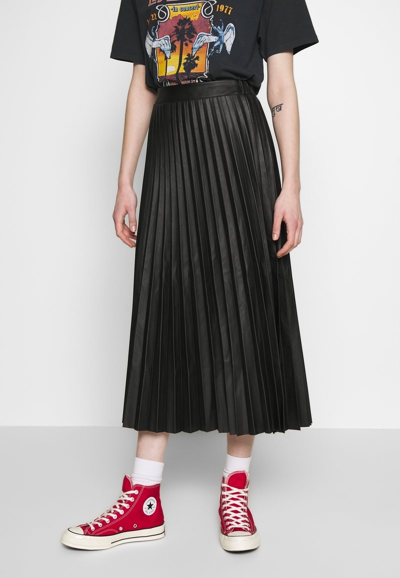 Personne portant un t-shirt graphique noir, une jupe midi plissée noire, des chaussettes blanches et des baskets montantes rouges sur un fond uni.