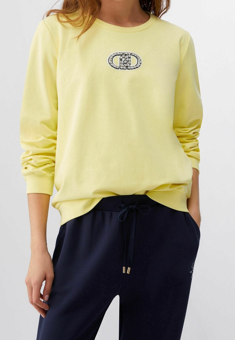 Femme portant un t-shirt jaune à manches longues avec un logo ovale noir et blanc sur la poitrine et un pantalon noir à cordon avec les mains dans les poches.
