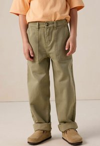 Olijfgroene, relaxed-fit cargobroek met voorplooien, zijzakken en opgerolde boorden, gecombineerd met een lichtoranje T-shirt met korte mouwen en bruine schoenen.