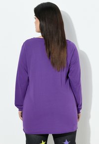 Kvinde med langt mørkt hår iført en ensfarvet lilla langærmet skjorte, stående med ryggen til mod en hvid væg.