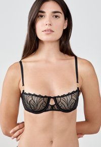 Soutien-gorge en dentelle noire avec des bonnets en maille transparente présentant un motif floral. Bords festonnés et fines bretelles réglables avec des détails en métal doré.