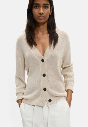 Kobieta o długich ciemnych włosach, ubrana w beżowy dziergany sweter z czterema czarnymi guzikami i białe spodnie, dłonie w kieszeniach, neutralne tło.