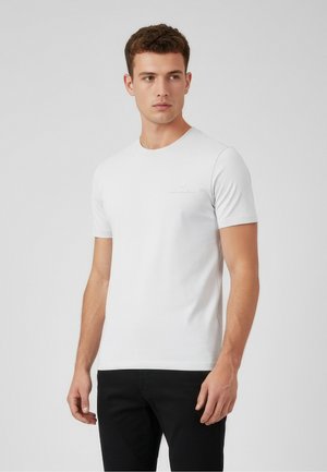 Jonge man die een eenvoudig wit t-shirt met korte mouwen en een zwarte broek draagt, staand tegen een neutrale grijze achtergrond.