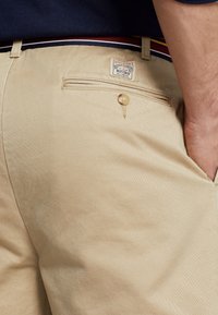 Chinos beige con una textura suave, que cuentan con un bolsillo trasero y cierre de botón. Se puede ver una etiqueta cerca de la cinturilla con un acento de rayas multicolores.