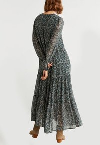 Langes grünes Blumenkleid aus transparenter Stoff, mit langen Ärmeln und einem gestuften Rock. Es hat kleine rosa und weiße Blumen und eine fließende Silhouette.