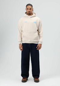 Beige hoodie met een voorzak, voorzien van blauwe logo's. Gecombineerd met losse donkere spijkerbroek en bruine laarzen met rode veters.