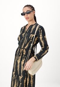 Juoda suknelė su auksiniu abstrakčiu raštu, ilgos rankovės, apykaklė, diržas liemeniui. Modelis laiko smėlinės spalvos per petį nešiojamą maišelį ir dėvi saulės akinius.