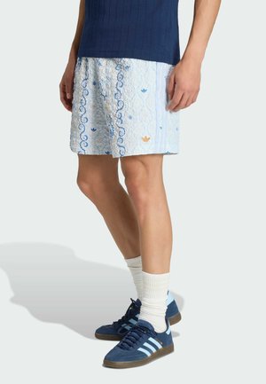 Mężczyzna w jasnoniebieskich, wzorzystych szortach, granatowej koszuli, białych skarpetkach oraz granatowych butach Adidas z jasnoniebieskimi paskami, stojący bokiem.