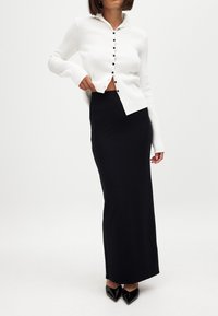 Bela rebrasta cardigan s gumbi, oblečena čez črno, tesno prilegajočo maxi krilo. Obleka vključuje elegantne črne špicate.