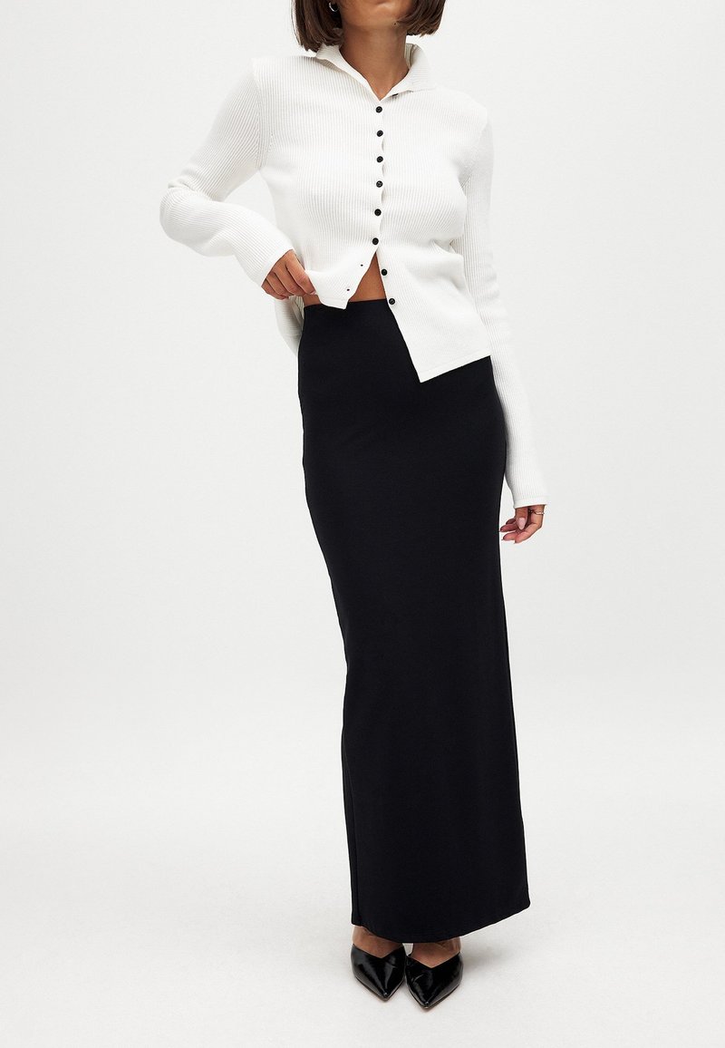 Bela rebrasta cardigan s gumbi, oblečena čez črno, tesno prilegajočo maxi krilo. Obleka vključuje elegantne črne špicate.