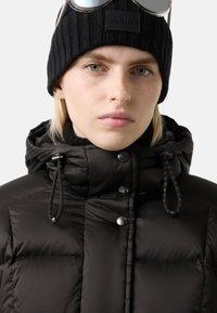 Černá péřová bunda na zip s vysokým límcem, knoflíkovými uzávěry a nastavitelnými šňůrkami. Oblékaná s černou žebrovanou pletenou čepicí.
