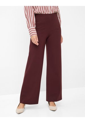 Frau trägt burgunderrote Hose mit weitem Bein, gestreiftes Langarmshirt und beige flache Schuhe, steht vor weißem Hintergrund.