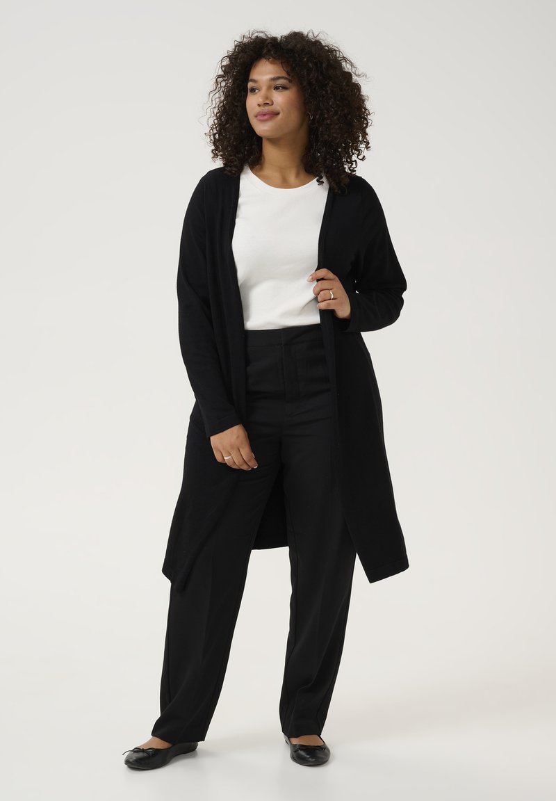 Zwarte lange cardigan met open voorkant, gedragen over een witte top en zwarte broek. Beschikt over een ontspannen pasvorm en zachte textuur. Een paar zwarte flats.