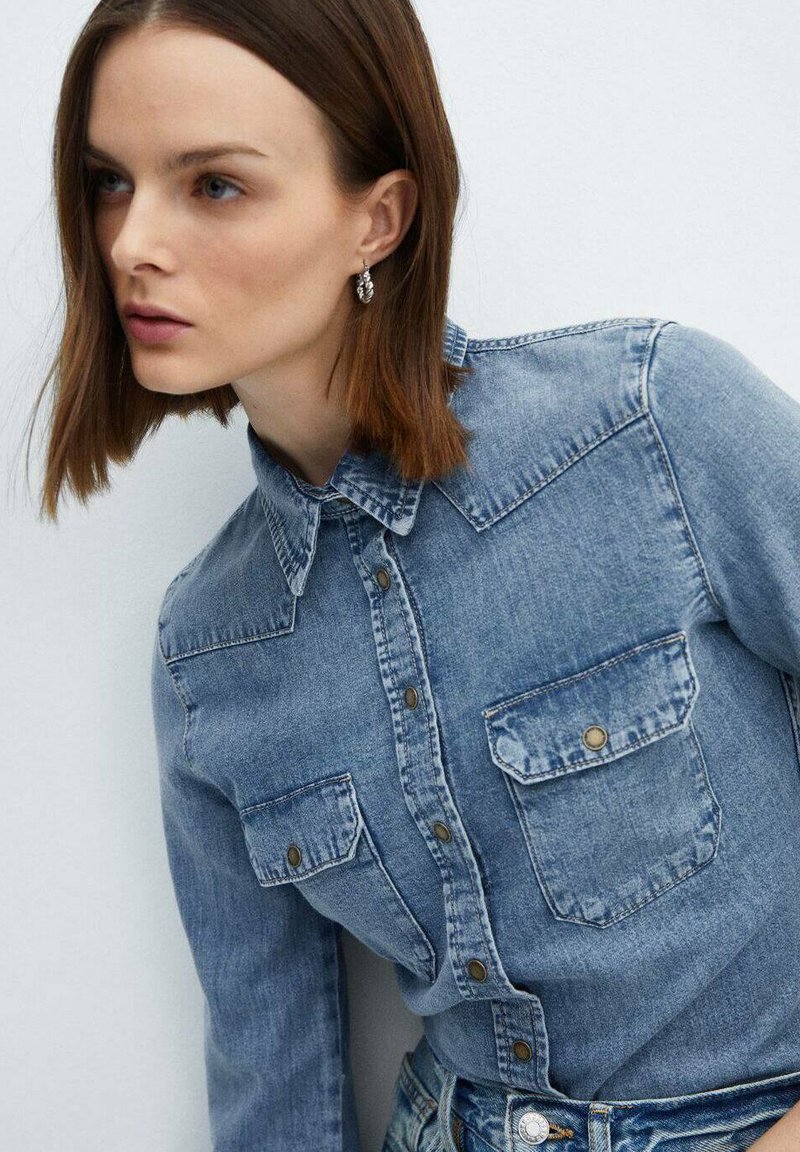 Hellblaues Jeanshemd mit durchgehender Knopfleiste, spitzem Kragen und zwei Fronttaschen. Verfügt über eine leicht verblichene Textur und metallische Knöpfe.