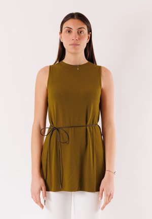 Donna con capelli lunghi e scuri che indossa un top verde oliva senza maniche con una sottile cintura nera annodata in vita, pantaloni bianchi, in piedi davanti a uno sfondo semplice.