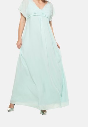 Femme portant une robe longue bleu clair fluide jusqu'au sol avec des manches courtes évasées et des escarpins argentés à bout pointu, sur un fond uni.