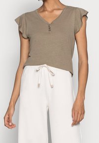 Donna che indossa una maglietta beige chiaro a V con bottoni e pantaloni bianchi con coulisse, mostrata dalla spalla fino a metà coscia.