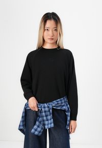 Jonge vrouw met lang blond haar draagt een zwarte sweater, een blauw geruit shirt geknoopt om haar middel, en donker wijde jeans, staande tegen een witte achtergrond.