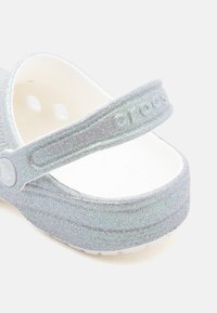 Srebrne błyszczące klapki Crocs z regulowanym paskiem na pięcie i otworami wentylacyjnymi na białym tle.