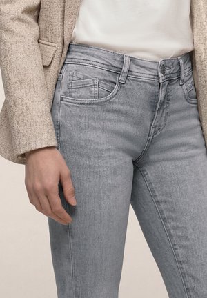 Osoba nosící světlé šedé džínové kalhoty, bílý top a béžové texturované sako s levou rukou volně podél těla.