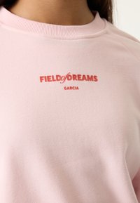 Primer plano de una sudadera rosa con texto rojo "FIELD of DREAMS" y "GARCIA" impreso en el pecho cerca del cuello.