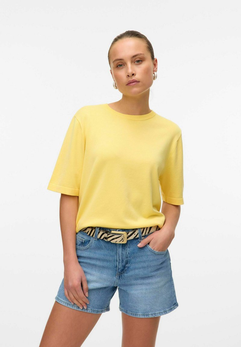 Gelbe Baumwoll T-shirt met korte mouwen, gecombineerd met blauwe denim shorts. Het model draagt een patroongenade band met een rechthoekige gesp.