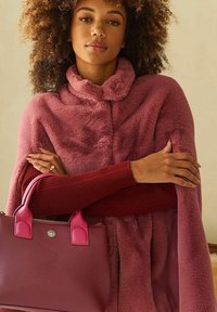 Roze nepbont poncho met een hoge kraag, gelaagd over een bordeauxrode geribde trui. Een bijpassende roze handtas met ronde handgrepen wordt vastgehouden.