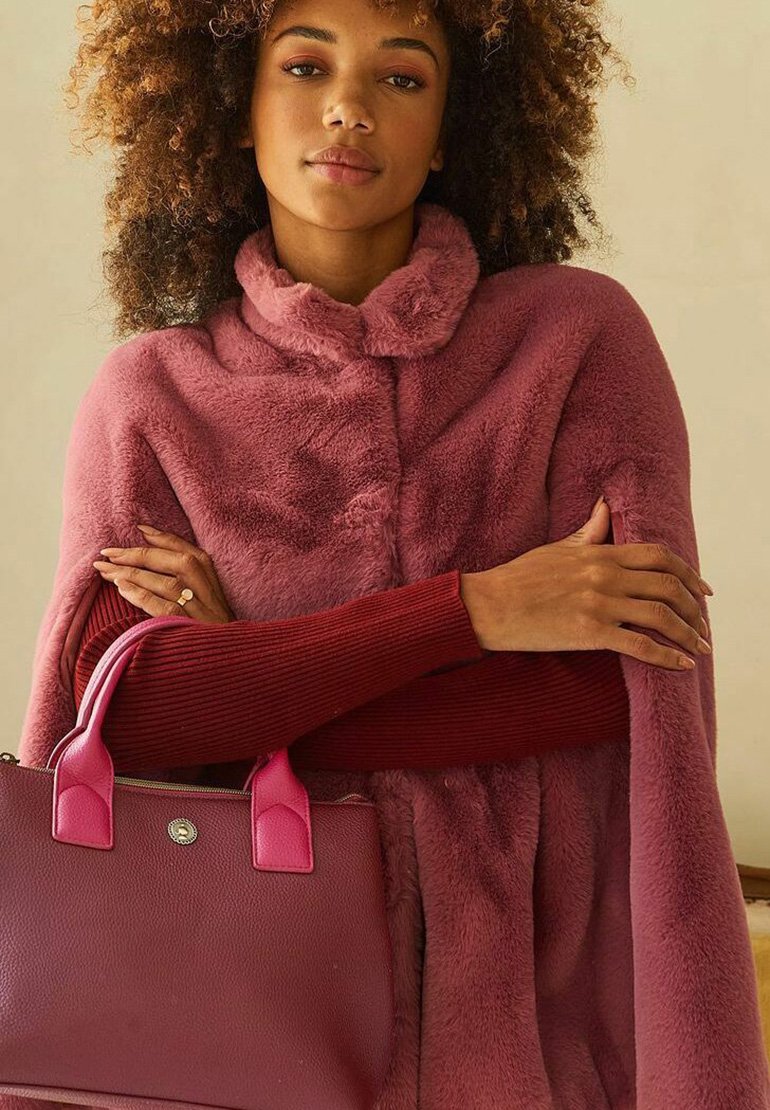 Roze nepbont poncho met een hoge kraag, gelaagd over een bordeauxrode geribde trui. Een bijpassende roze handtas met ronde handgrepen wordt vastgehouden.
