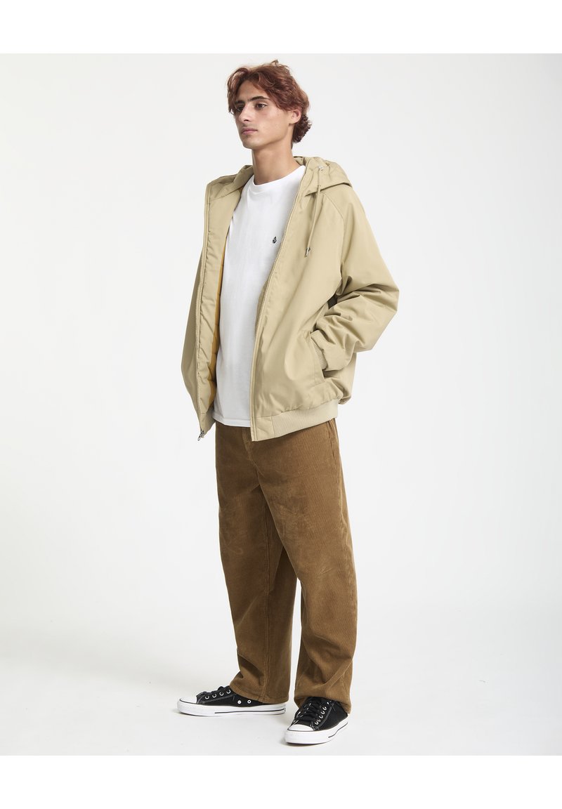 Giacca beige con cappuccio e chiusura con zip, indossata sopra una t-shirt bianca, abbinata a pantaloni di velluto a coste marroni e sneakers nere. Design semplice.