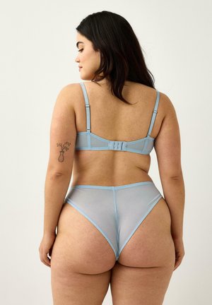 Kurvet kvinde med mørkt hår iført lyseblå gennemsigtig bh og trusser med høj talje, der viser en blomstertatovering på hendes venstre overarm.