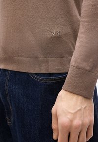 Gros plan sur le torse d'une personne portant un pull marron avec une broderie "MN" et un jean bleu foncé, la main détendue sur le côté.