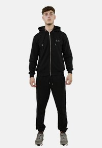 Felpa nera con zip, tasca frontale, polsini a costine e cappuccio; abbinata a pantaloni jogger neri, entrambi realizzati in tessuto morbido.