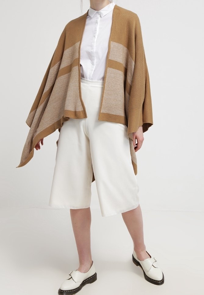 Bruin en beige gestreepte gebreide poncho met open front, gecombineerd met witte wijde shorts en zwarte schoenen. Gladde textuur en comfortabele pasvorm.