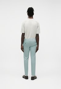 Άνδρας με σκουρόχρωμο δέρμα και πλεξούδες στέκεται με την πλάτη προς τα εμπρός, φορώντας ανοιχτό γκρι κοντομάνικο πουκάμισο, ανοιχτό μπλε παντελόνι και σκοτεινά παπούτσια.