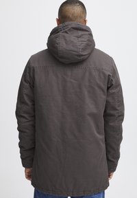 Chaqueta marrón con capucha, de textura suave, que presenta un corte holgado, costuras visibles y un dobladillo trasero más largo.