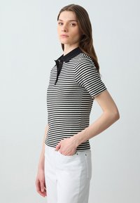 Polo a righe bianche e nere con maniche corte, tessuto strutturato e colletto nero a tinta unita. Indossato con pantaloni bianchi.