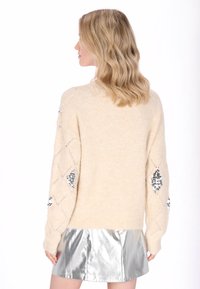 Pulover bej tricotat cu accente din paiete pe mâneci, având croială lejeră și manșete striate, asortat cu o fustă mini metalizată argintie.