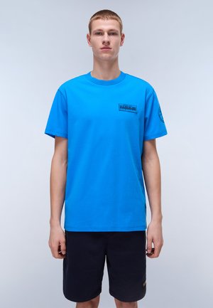 Blå bomulds T-shirt med korte ærmer, rund halsudskæring og et lille sort logo trykt på brystet. Båret med mørke shorts.
