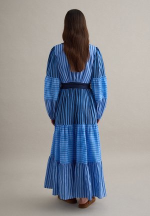 Vrouw met lang bruin haar draagt een lange blauwe patchworkjurk met afwisselende horizontale en verticale strepen, staand tegen een effen achtergrond.