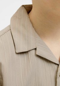 Bežinė maršina su tekstūruota medžiaga, vertikaliomis juostelėmis, įpjautu apykakle ir sagčių užsegimu priekyje. Detalus apykaklės ir kaklo vaizdas.