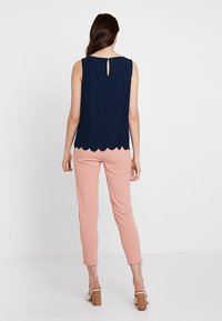 Top sin mangas color marino con dobladillo festoneado y detalle de abertura en la espalda, combinado con pantalones ajustados color rosa claro y zapatos de tacón cuadrado marrones.