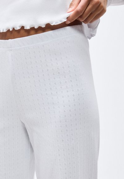 Κοντινό πλάνο ενός ατόμου που σηκώνει ένα λευκό, ριγωτό πουκάμισο με σχέδιο ματιών πάνω από αντίστοιχο ψηλόμεσο κολάν.