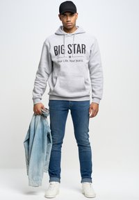 Šedá mikina s potiskem "BIG STAR", modré džínové kalhoty a bílé tenisky. Drží světle modrou džínovou bundu. Jednoduchý, ležérní outfit.