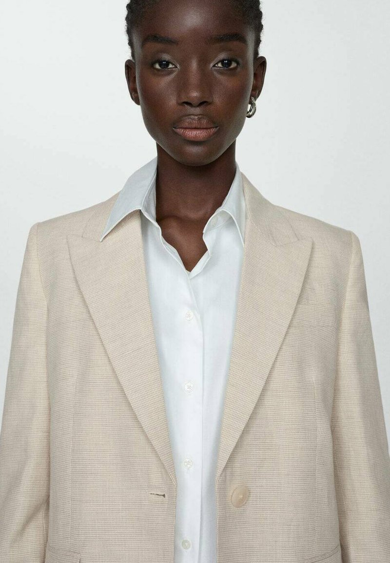 Beige karierter Blazer mit Reverskragen, Ein-Knopf-Verschluss, getragen über einem weißen Hemd mit glatter Textur.