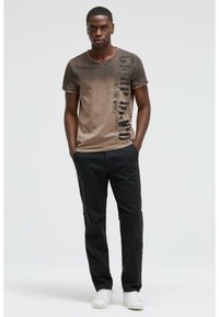 Brown gradient t-shirt with a v-neck, featuring vertical black text pattern, paired with black pants and white sneakers.