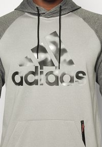 Sweat à capuche gris clair avec manches accentuées noires, arborant un grand logo Adidas noir et argent, cordon de serrage ajustable, et poche zippée.