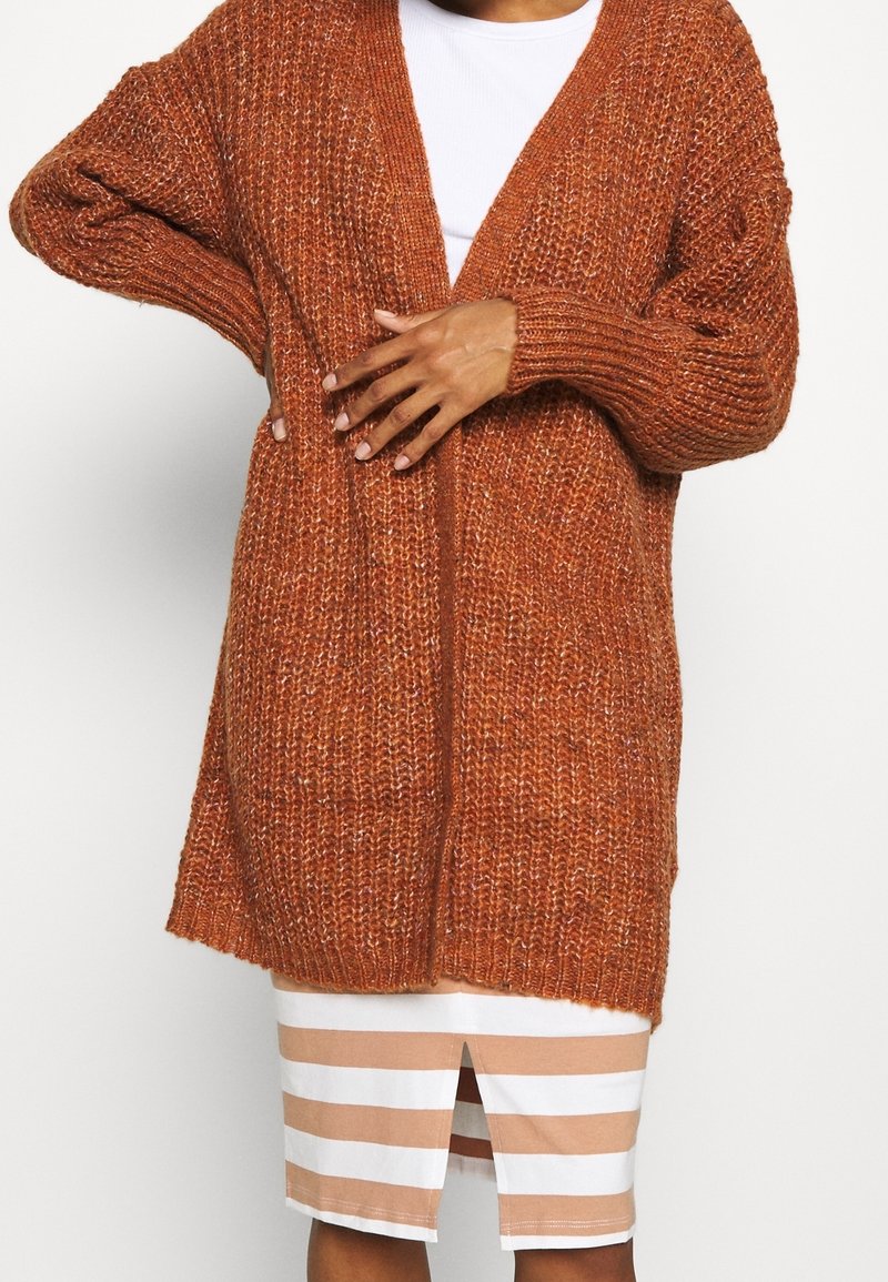 Chunky gebreid vest in verbrand oranje met een losse pasvorm, open voorkant en geribde mouwen. Gecombineerd met een wit top en gestreepte kokerrok.