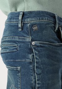 Faded blue denim jeans with a metal logo tag. Features stitched back pockets and a textured waistband.