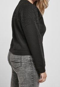 Femme portant un haut noir à manches longues avec épaules en dentelle et jean gris délavé, vue de profil sur un fond uni.
